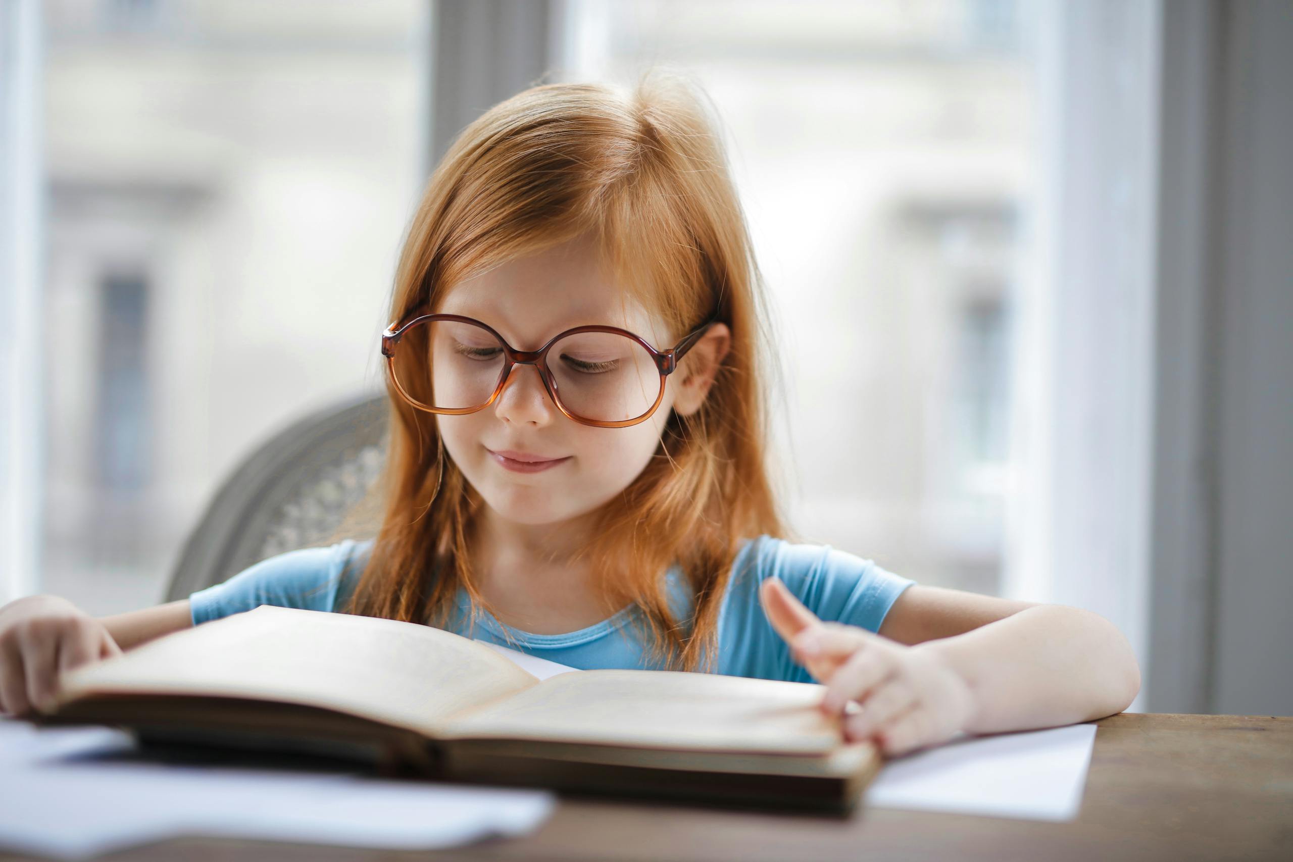 Charming young girl wearing oversized glasses reading book at home