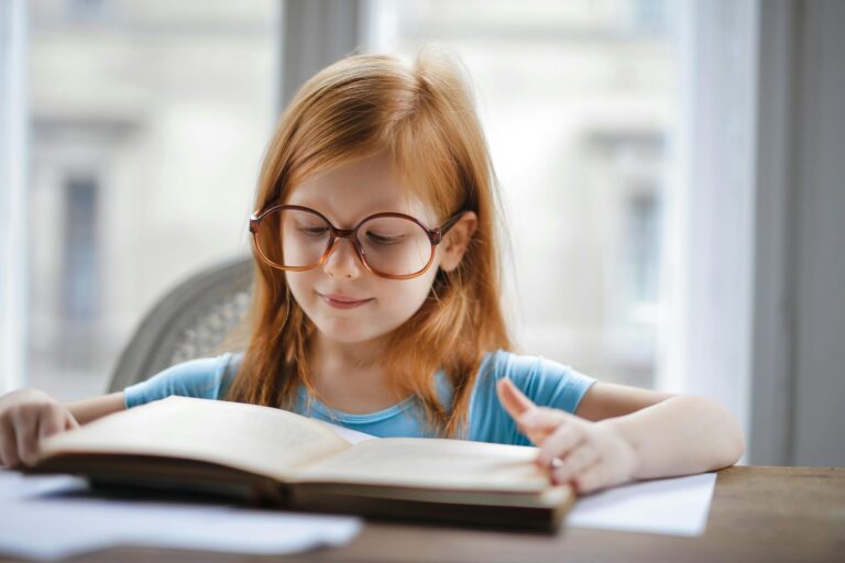 Charming young girl wearing oversized glasses reading book at home