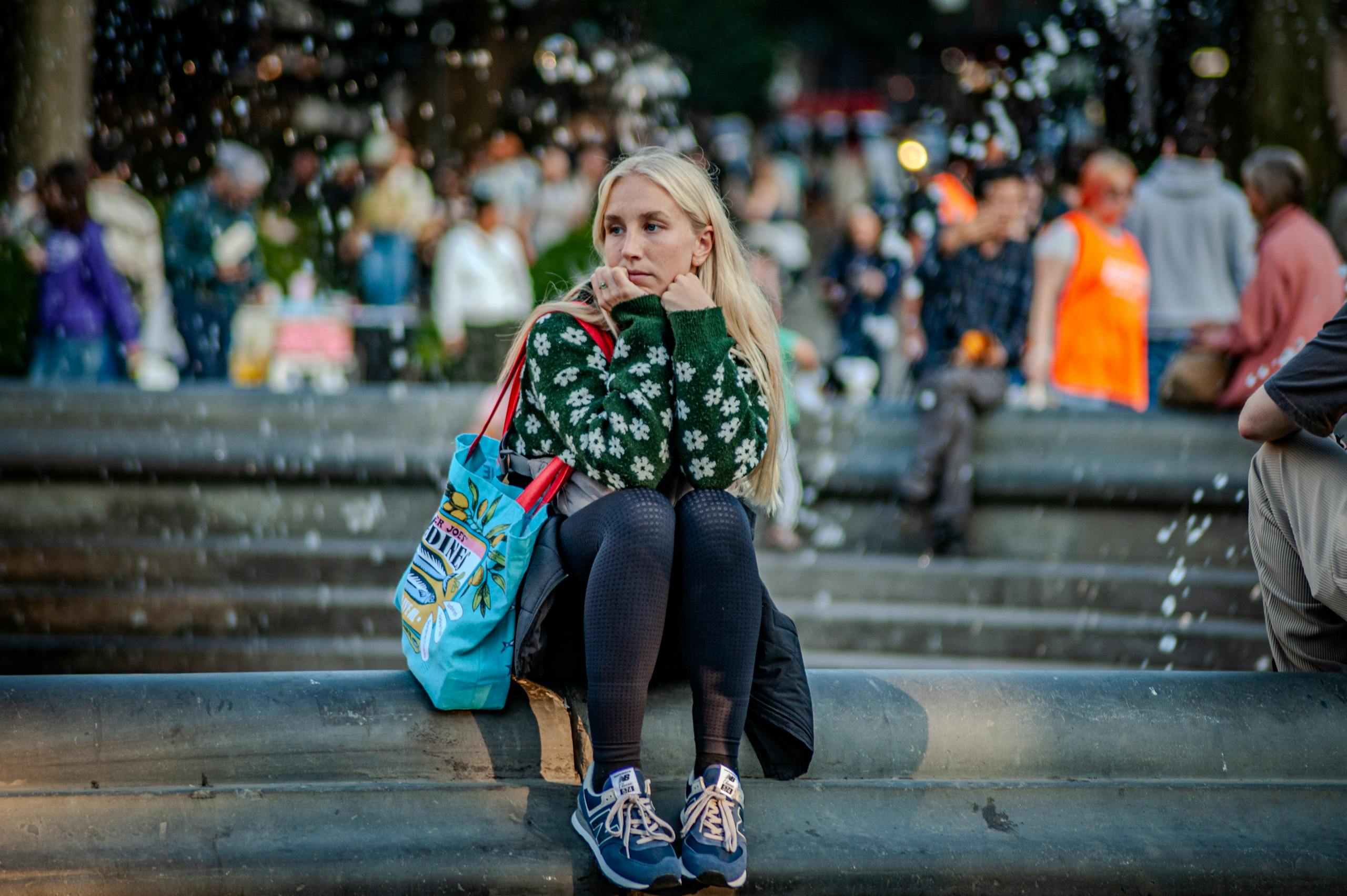 Contemplative woman sitting by fountain in busy park surrounded by crowds.