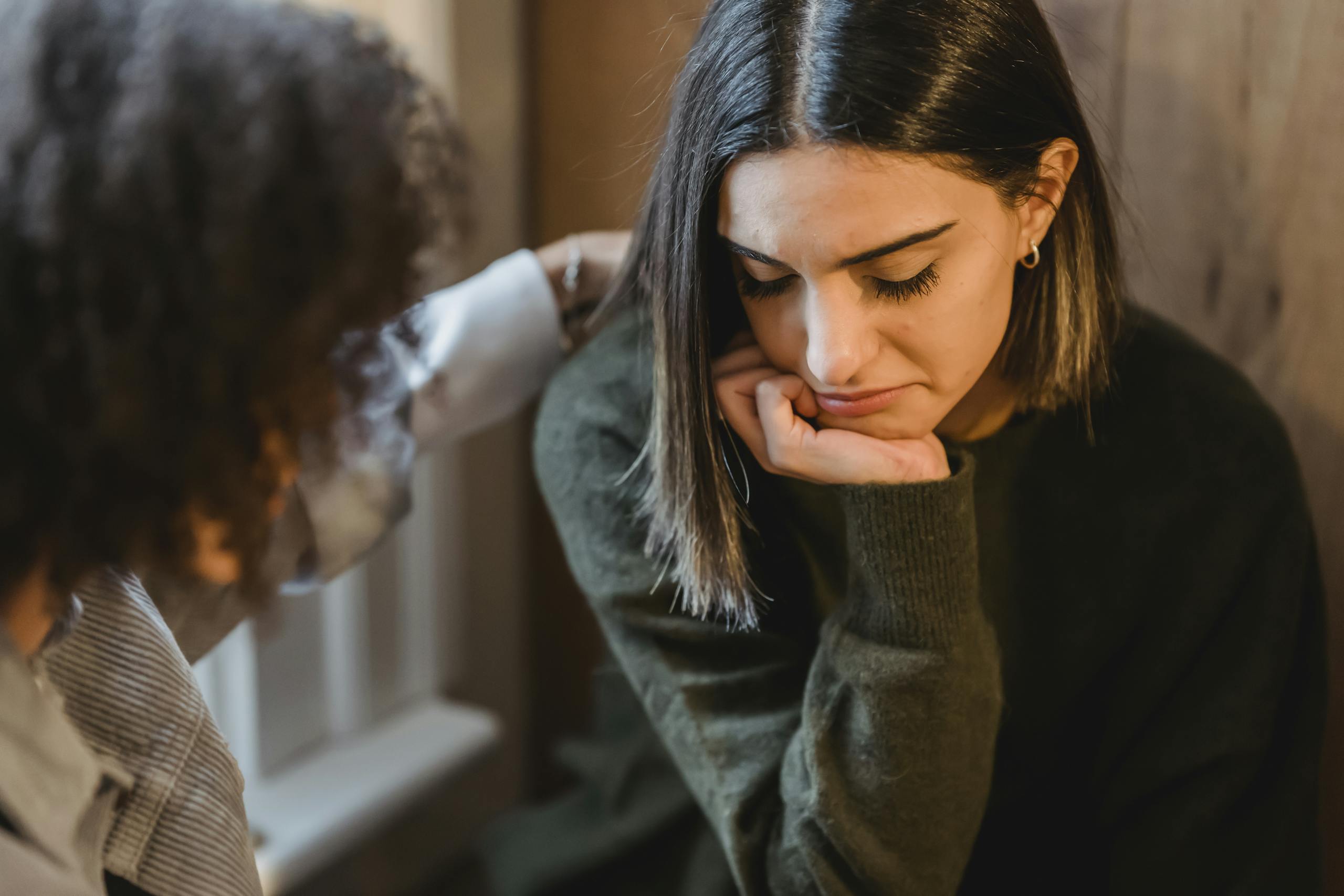 Woman comforting and tapping shoulder of upset friend while sitting together at home