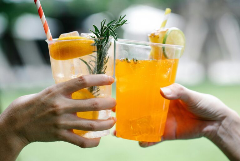 Two hands toasting with colorful citrus cocktails outdoors, rosemary garnish
