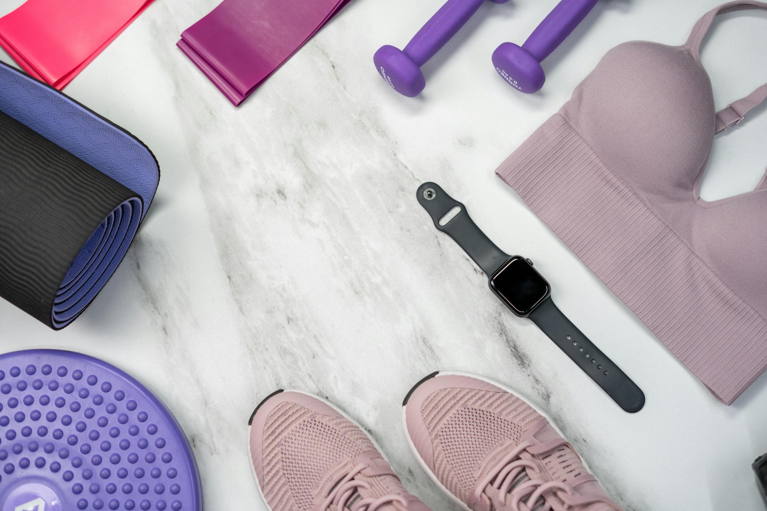 Flat lay of fitness equipment including yoga mat, dumbbells, and smartwatch on marble.