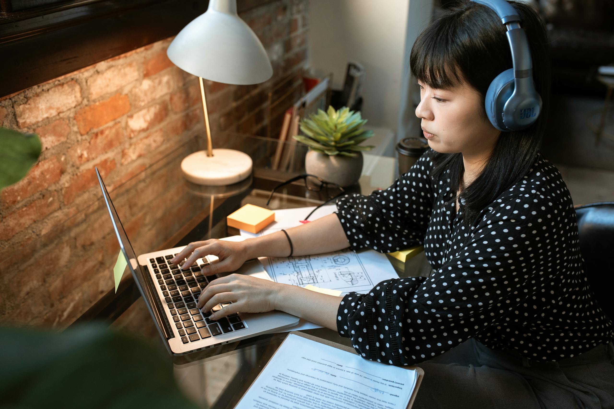 Focused woman wearing headphones working on laptop in cozy home office with natural light.