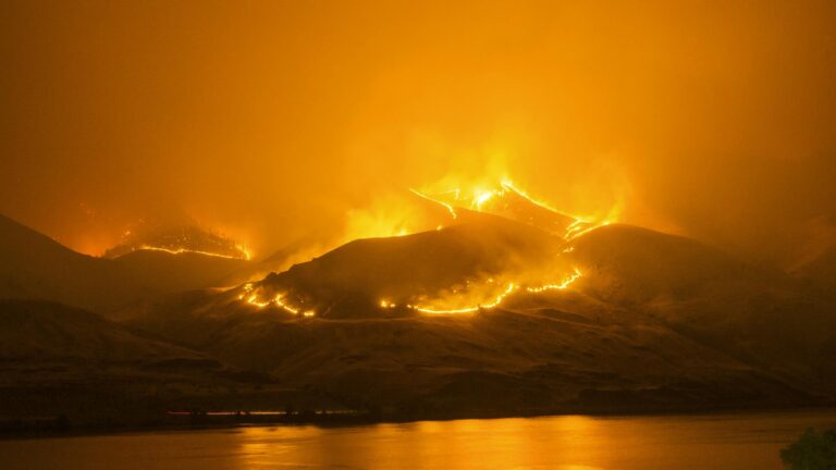Intense wildfire blazing on hills reflected in calm river below.