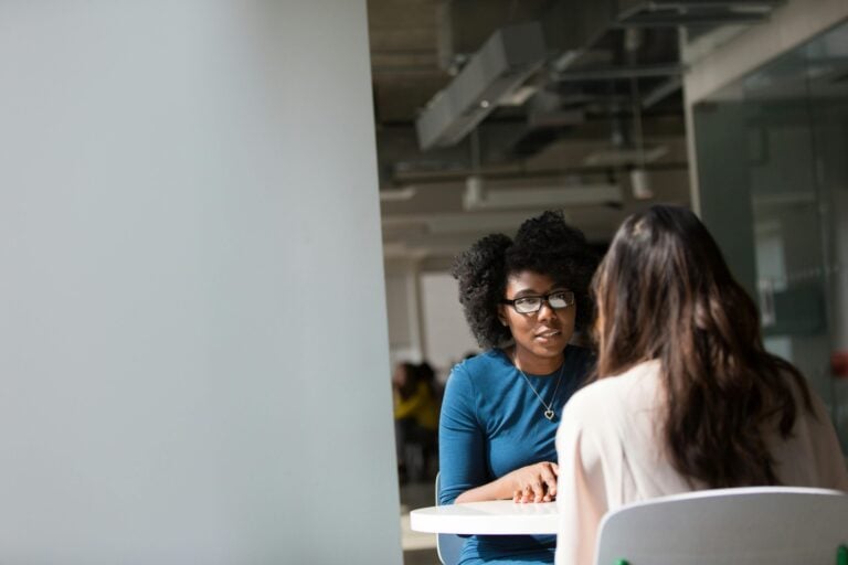 Two women having a focused one-on-one conversation at a table in a modern office setting.