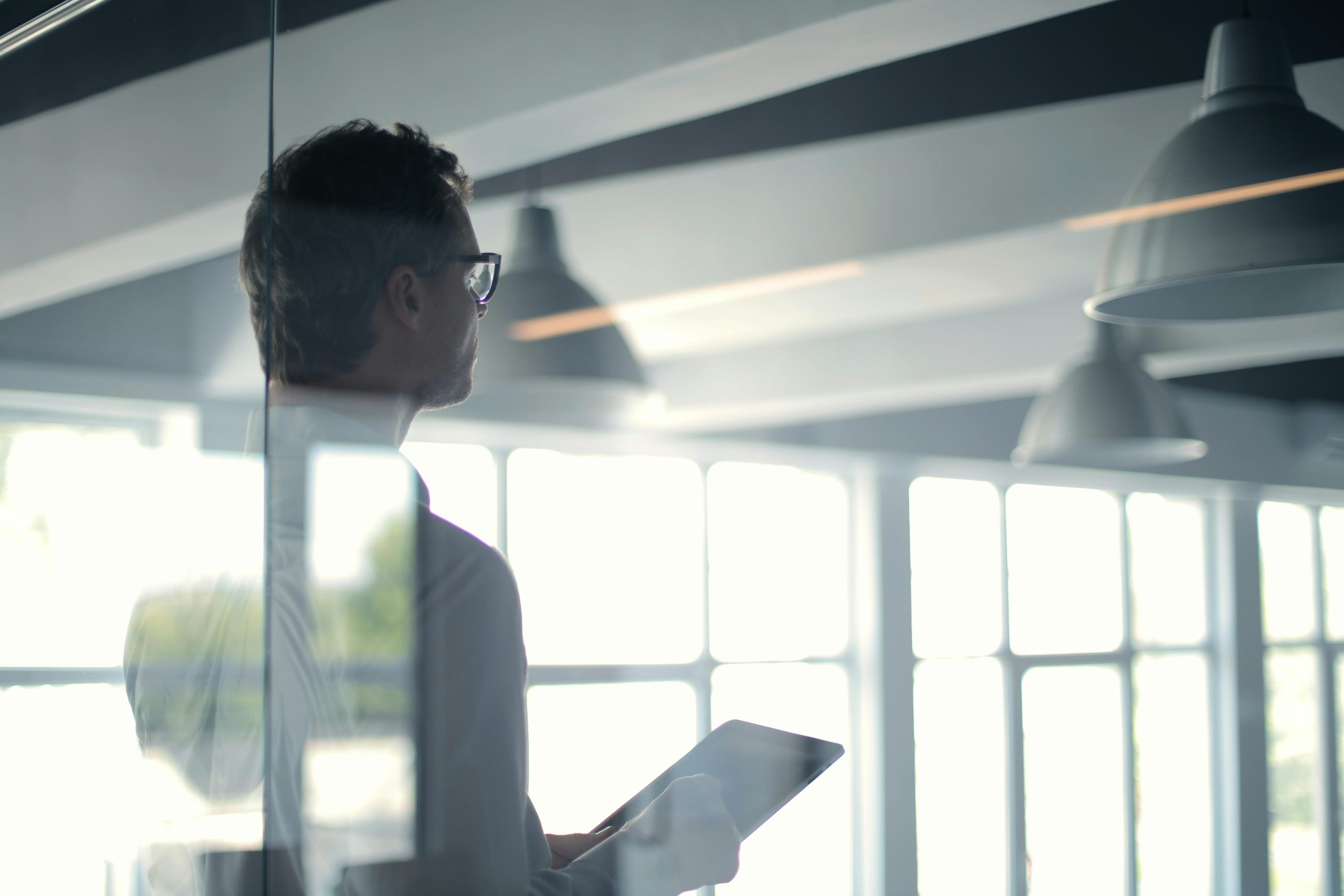 Thoughtful man with glasses holding tablet, gazing out bright windows alone