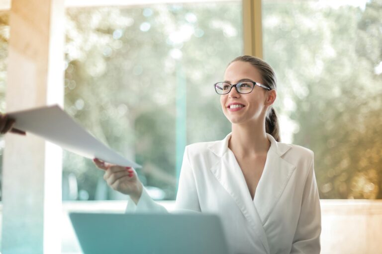 Female executive manager in professional attire passing documents to colleague at laptop