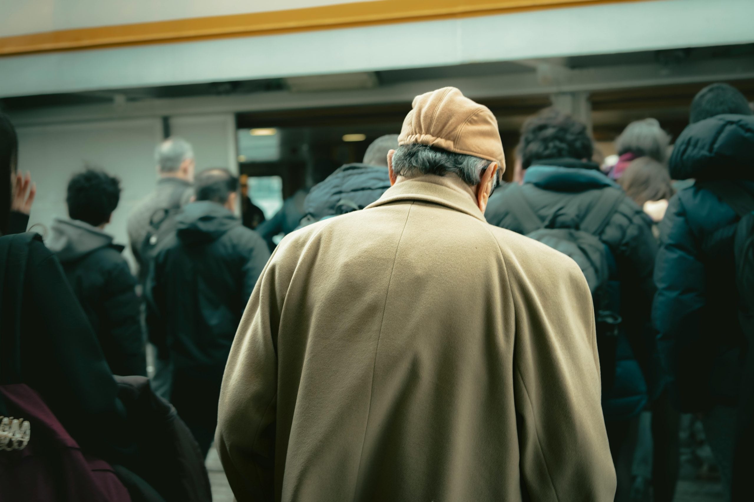 Solitary man in beige coat stands apart from crowded group of people indoors
