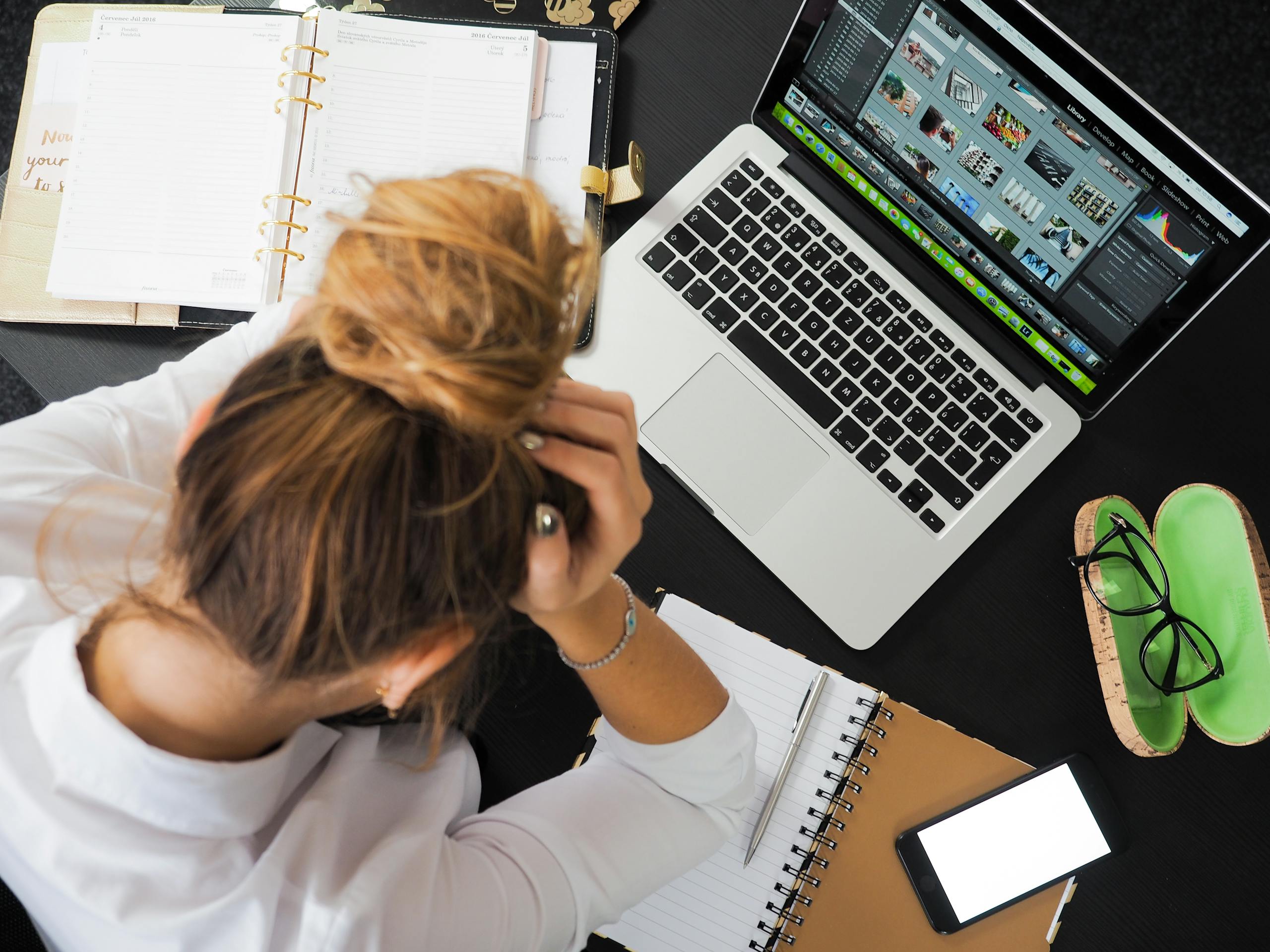 Overhead view of stressed woman at desk with laptop, phone, notebooks.