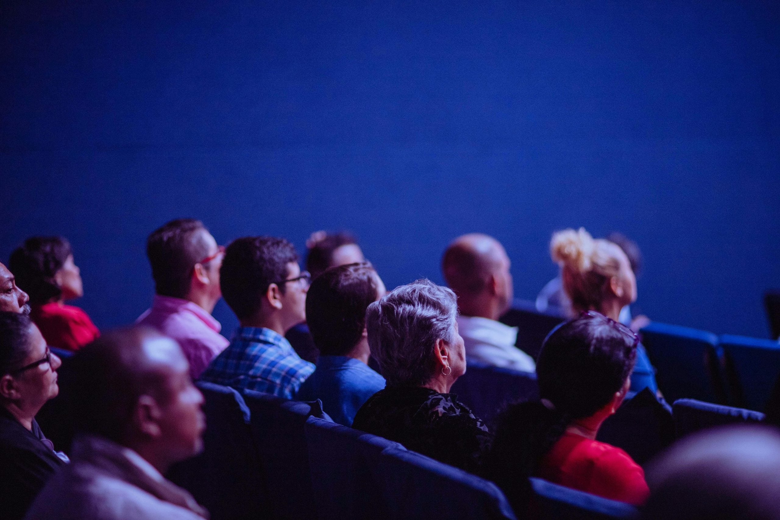 Group of diverse people sitting together in blue-lit theater, watching performance