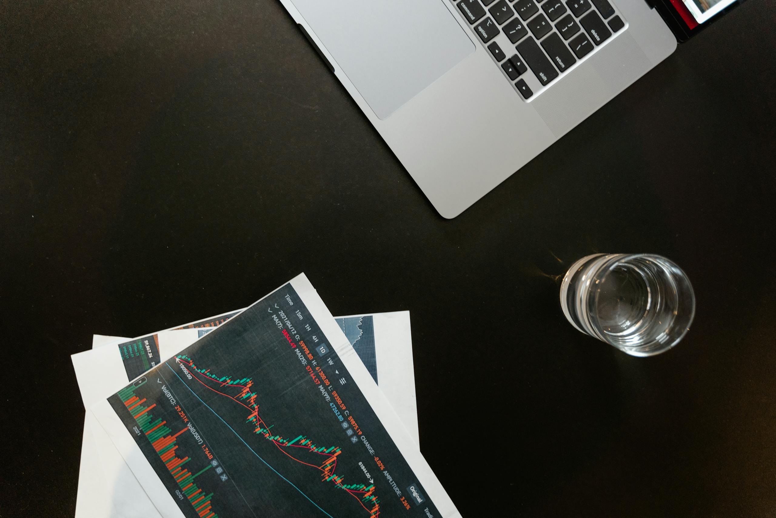 Professional workspace featuring financial graphs, laptop and water on desk.