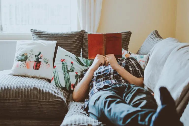 Relaxed person lying on sofa reading book surrounded by soft cushions.