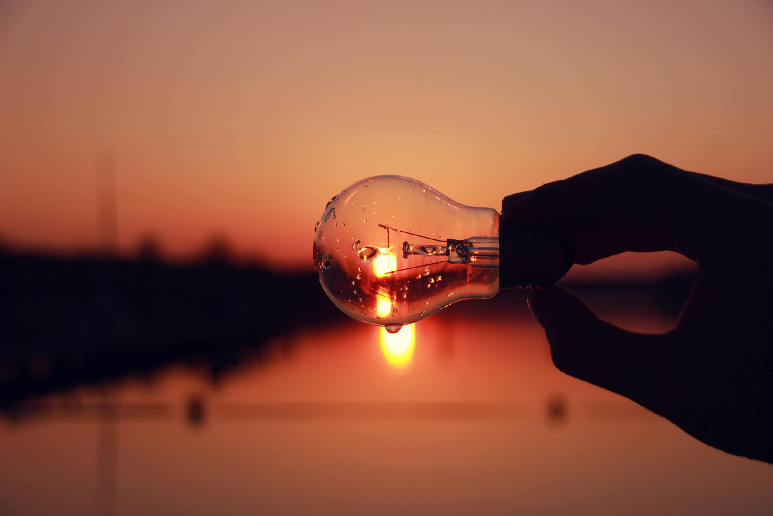 Silhouette of hand holding light bulb against stunning sunset over lake blending creativity and nature.