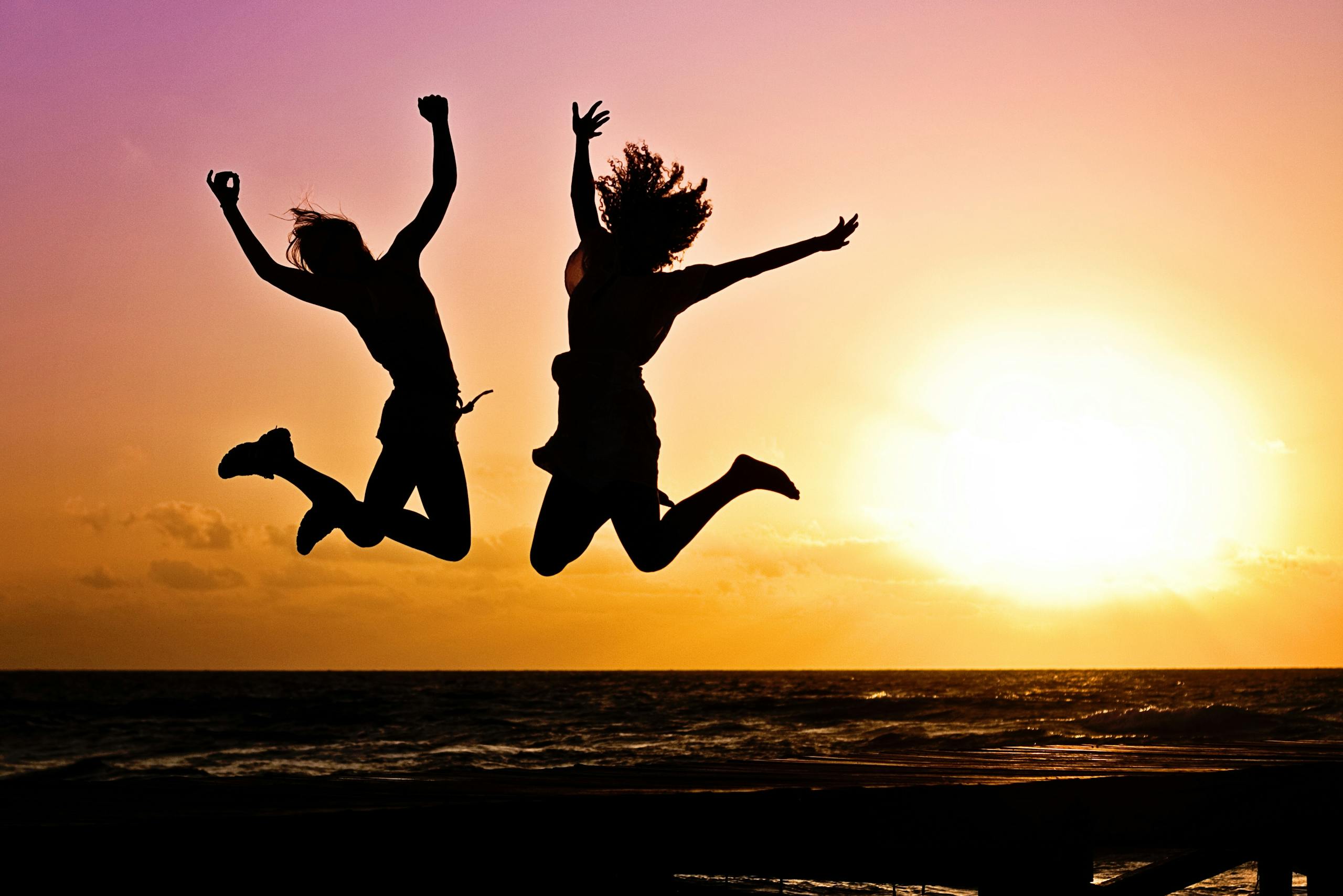 Silhouettes of two people joyfully jumping on beach during vibrant sunset.