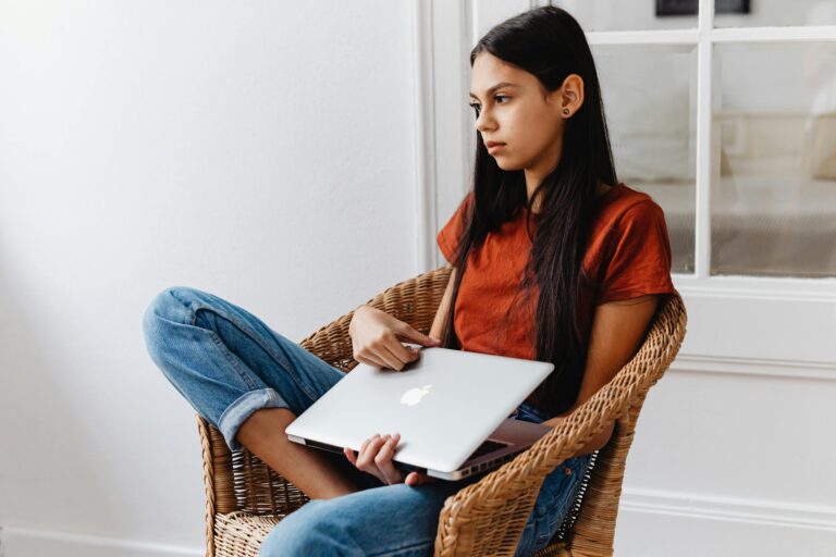 Teen girl lounging indoors in wicker chair using laptop