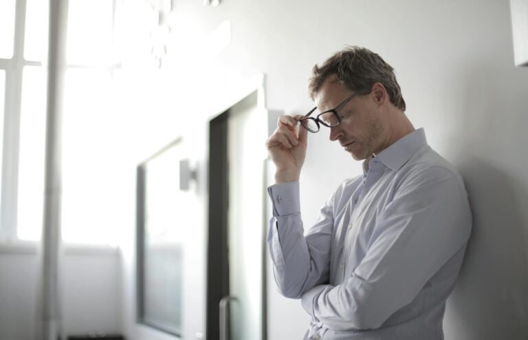 Thoughtful man in bright room holding glasses against wall contemplating