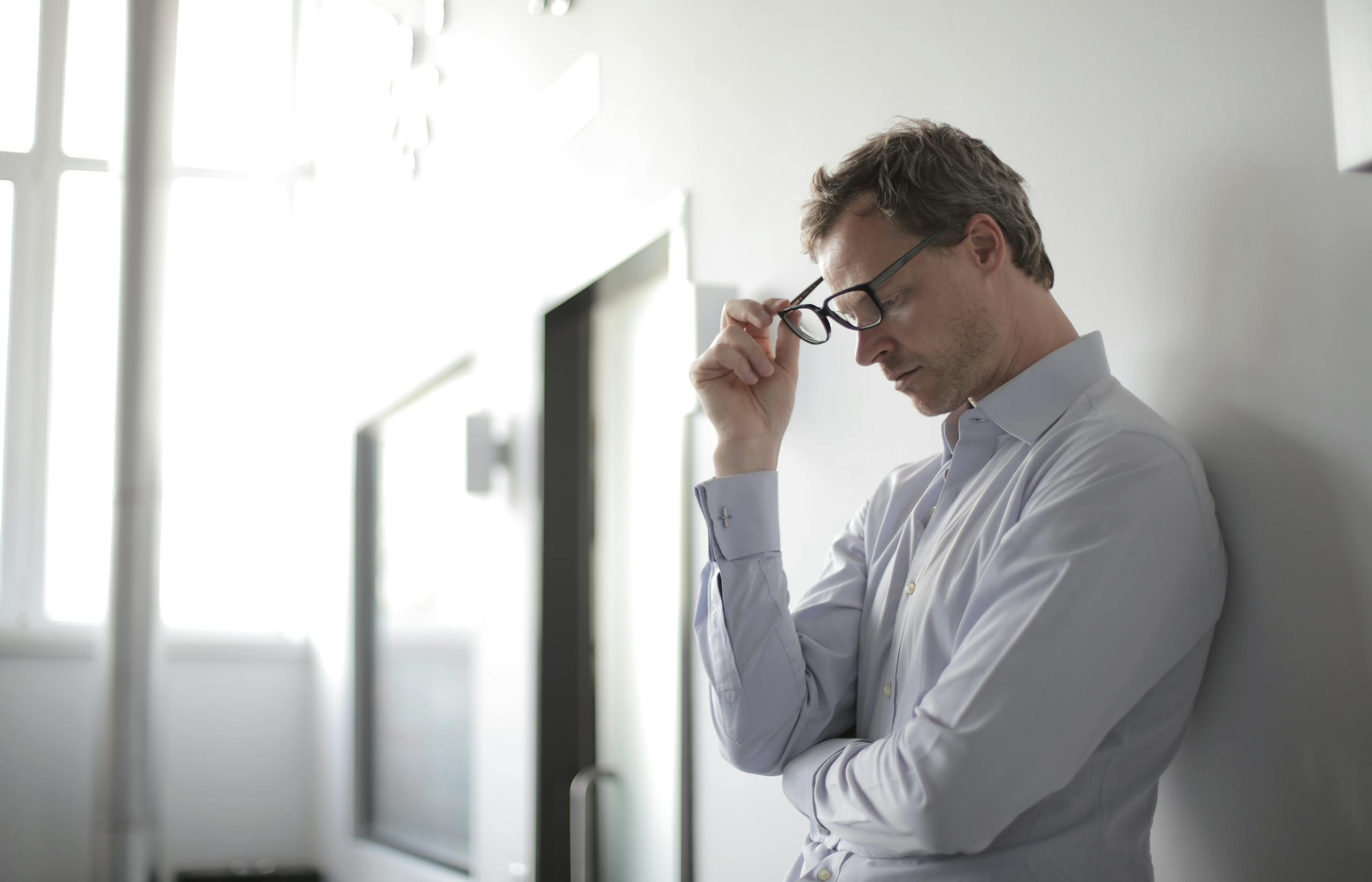 Thoughtful man in bright room holding glasses against wall contemplating