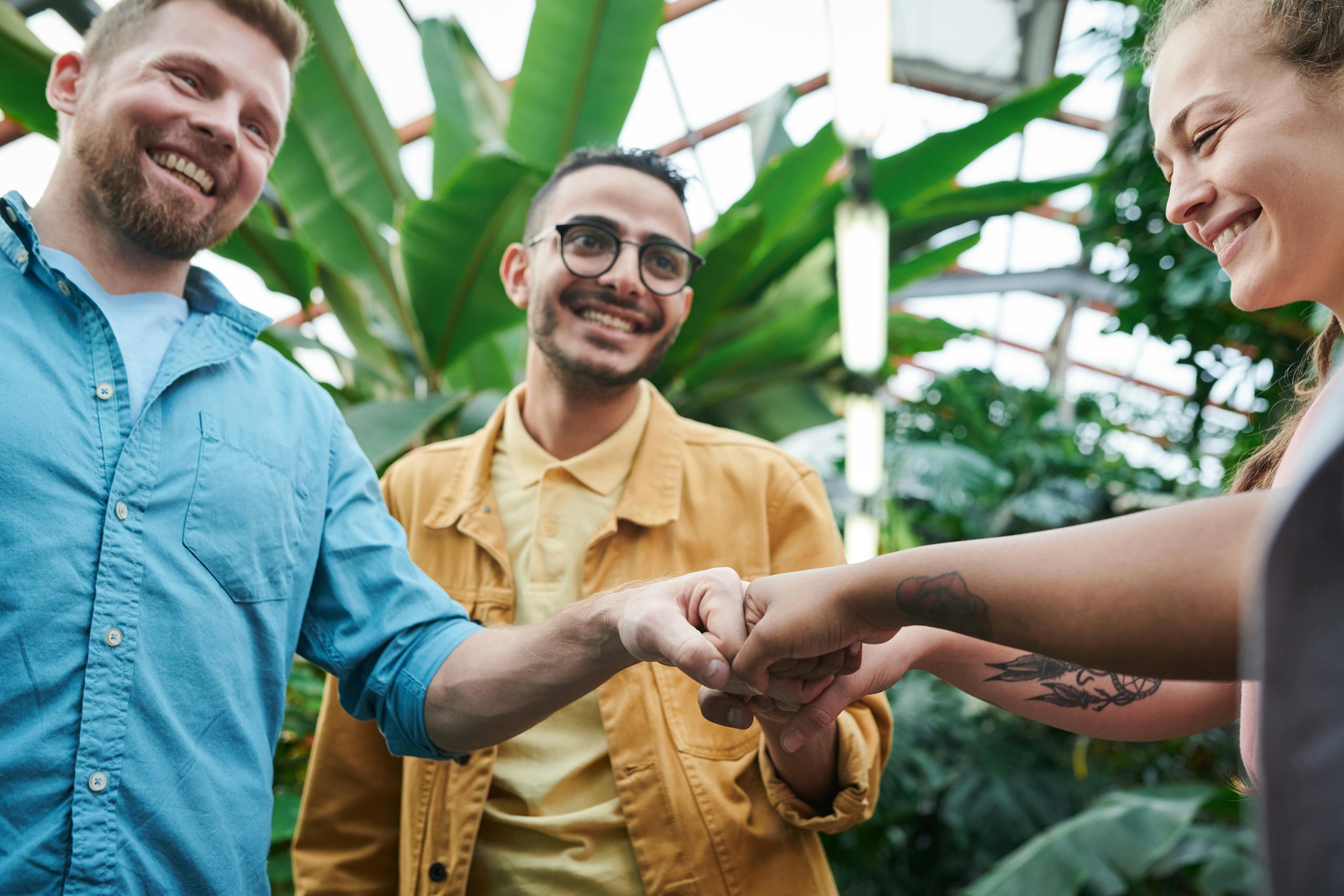 Three friends sharing joyful fist bump in lush indoor greenhouse environment