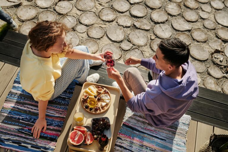 Overhead view of couple clinking wine glasses at sunny outdoor picnic with fruits.