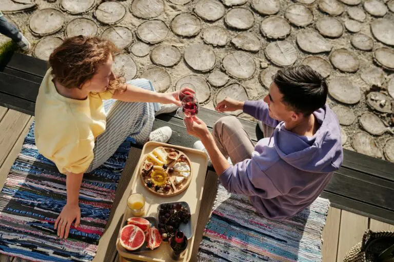 Overhead view of couple clinking wine glasses at sunny outdoor picnic with fruits.