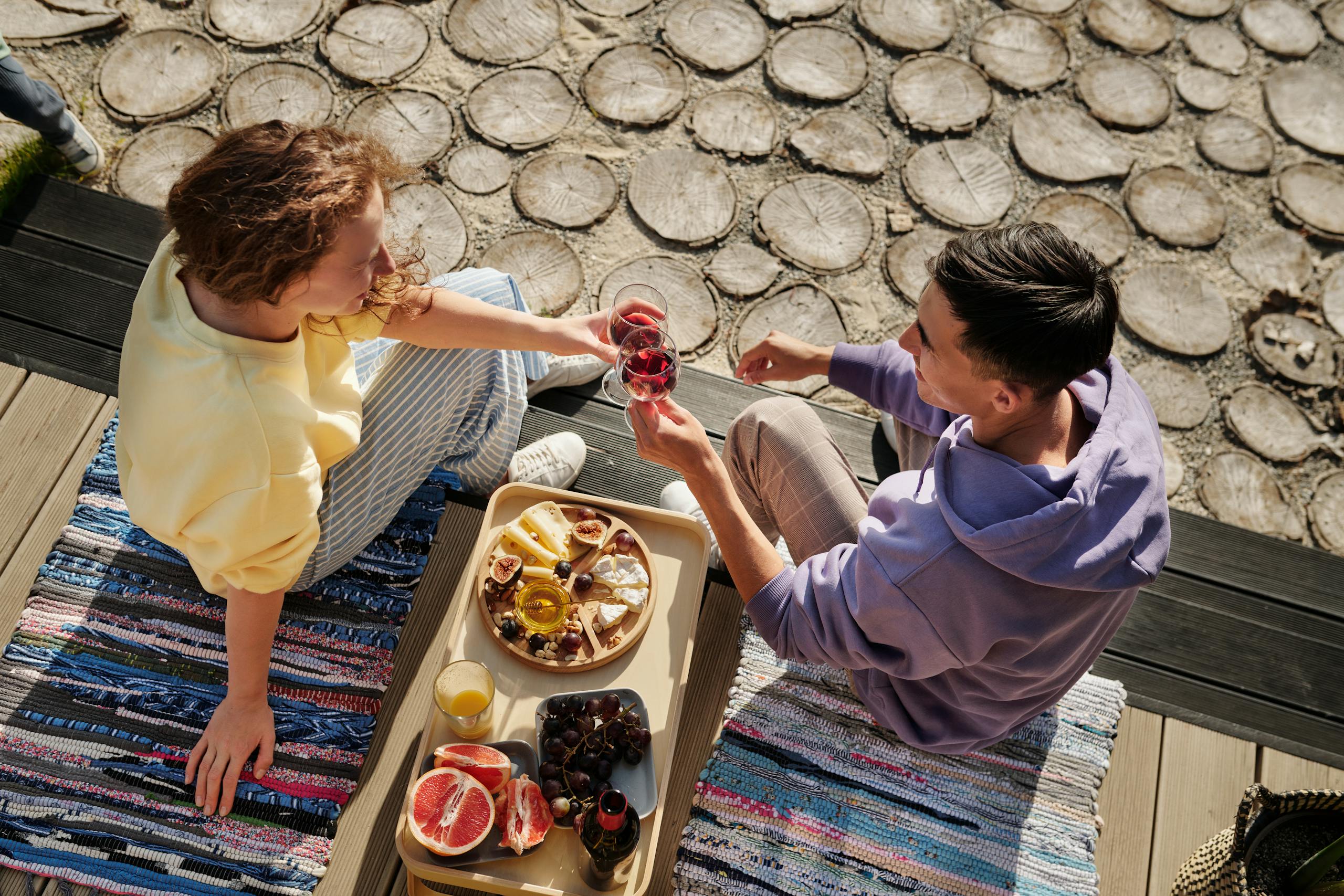 Overhead view of couple clinking wine glasses at sunny outdoor picnic with fruits.