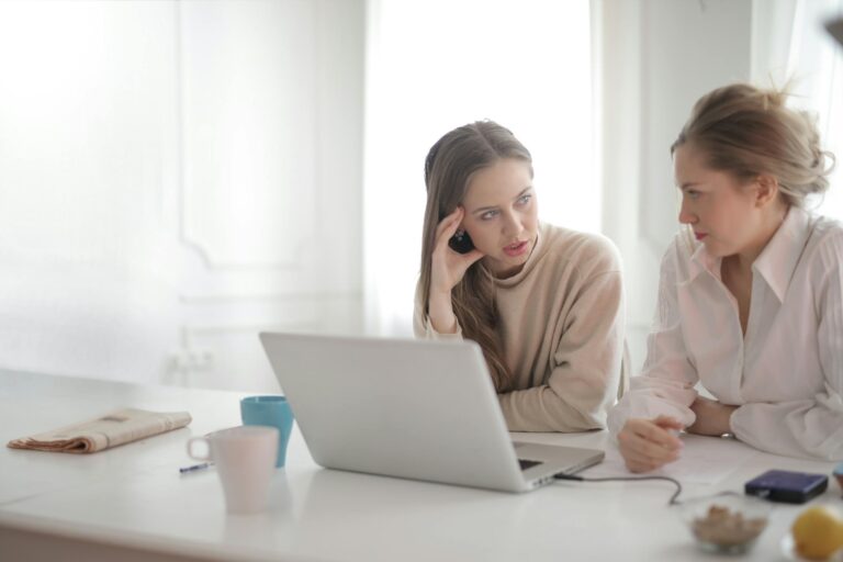 Two women engaged in focused work discussion at desk with laptop in bright office.
