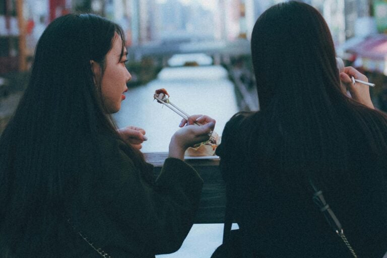 Two women enjoying meal by riverside with blurred urban background scenery.