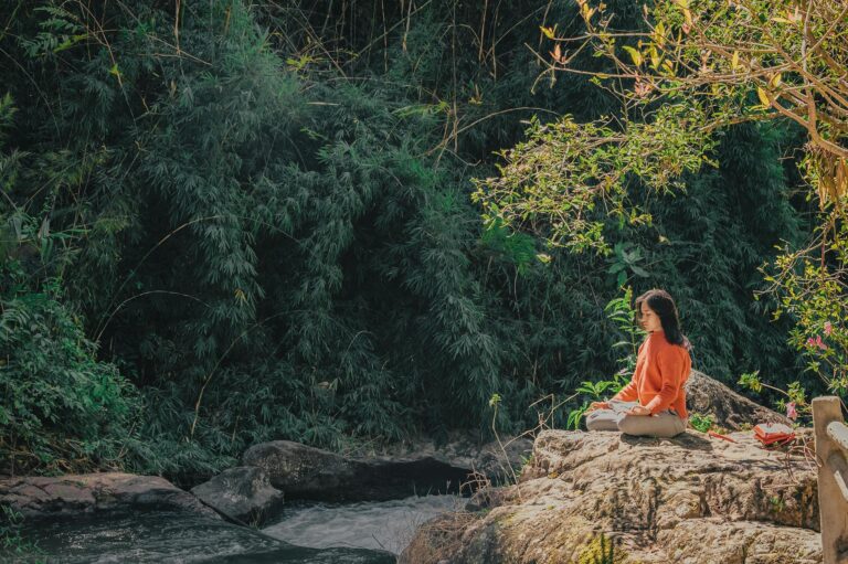 Woman meditating peacefully by riverside surrounded by lush greenery and sunshine.