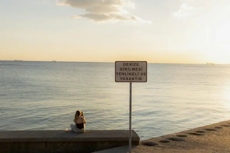 Woman sitting by the sea at sunset in Istanbul with water entry warning sign visible.