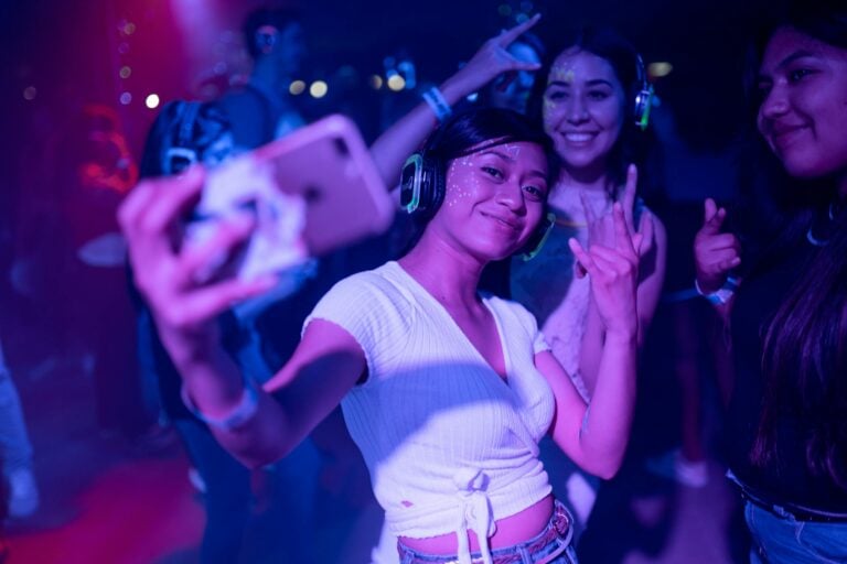 Young adults at silent disco party wearing headphones capturing selfies amid colorful lights.