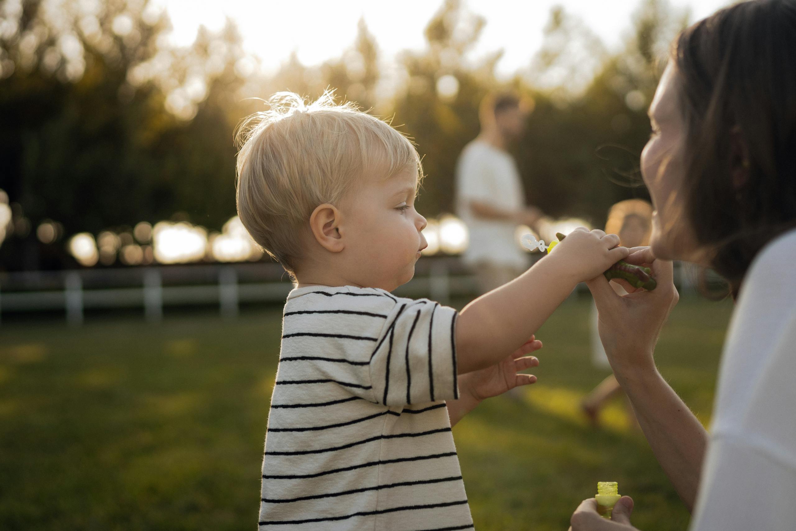 Young child blowing bubbles with adult in sunny park setting, playful moment.