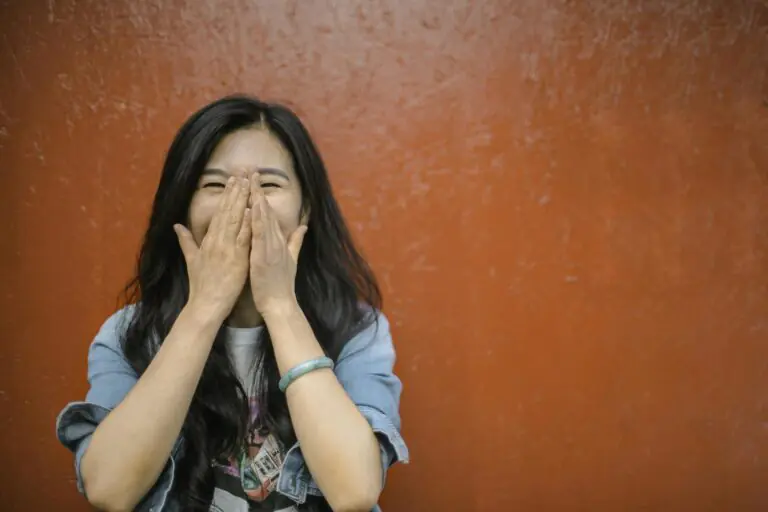Young Asian woman in casual outfit covering face with hands laughing against orange wall.