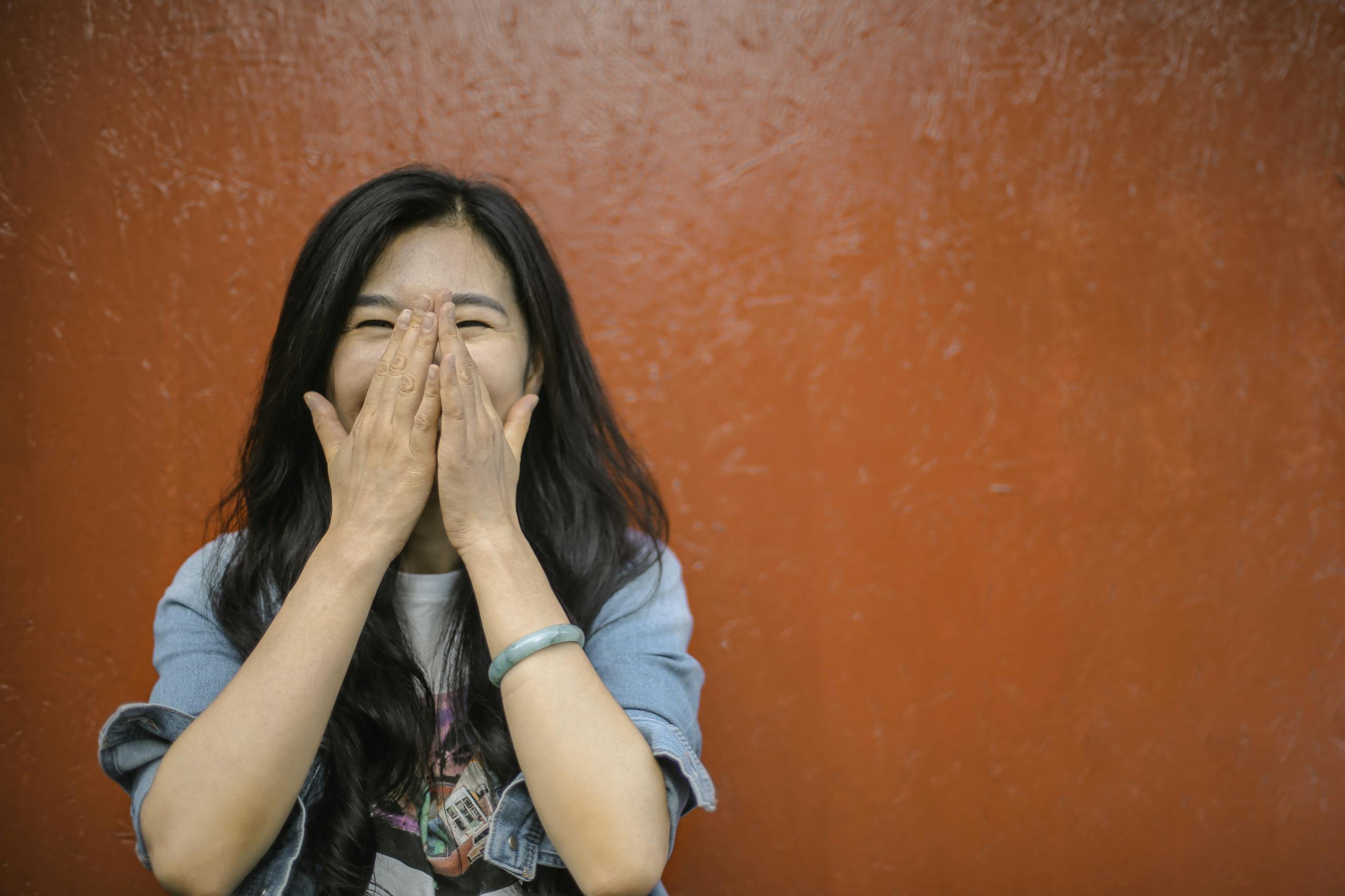Young Asian woman in casual outfit covering face with hands laughing against orange wall.