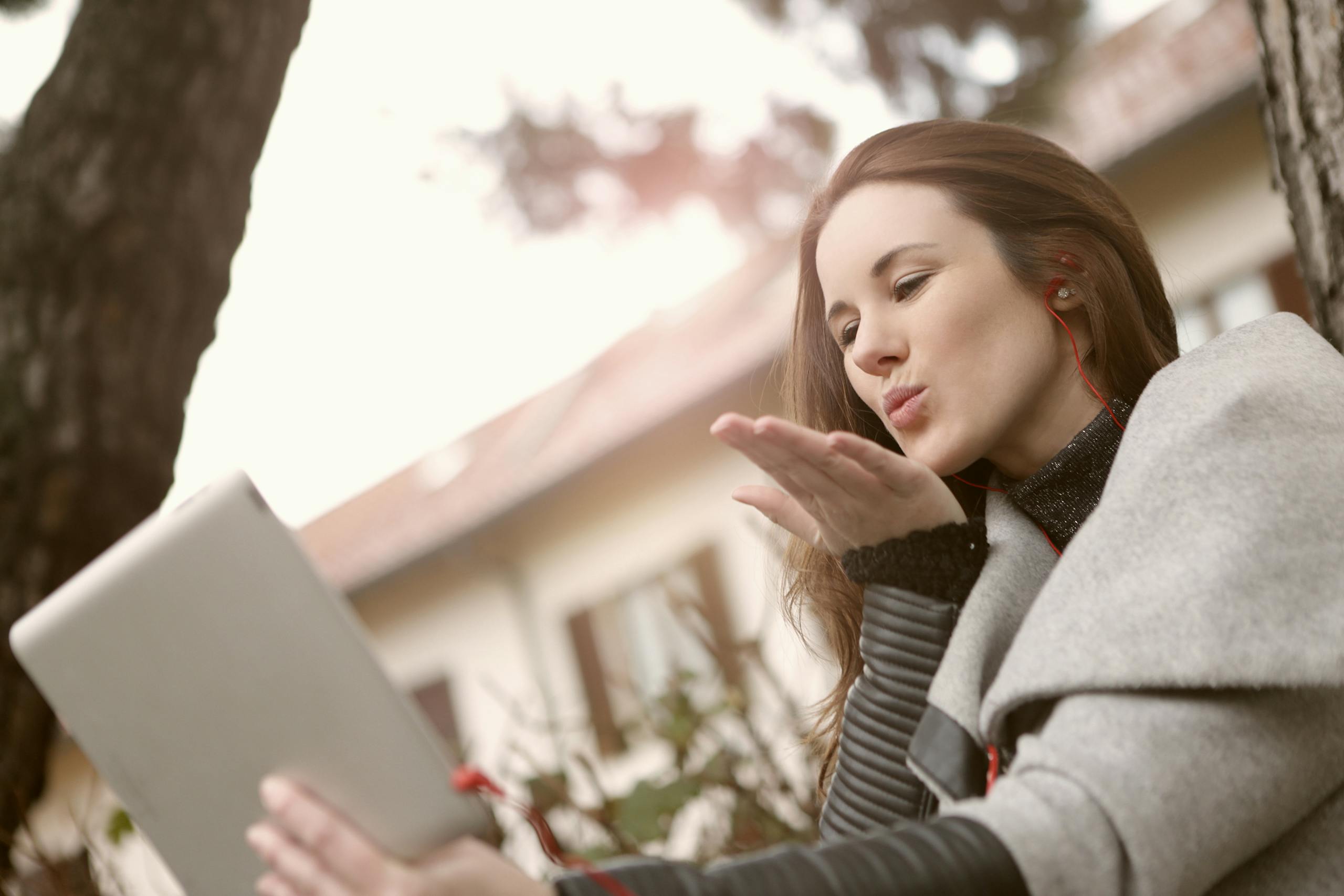 Young woman video calling outdoors on tablet with playful flying kiss gesture.