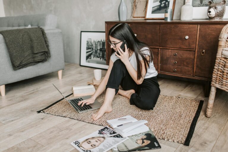 Young woman working from home on laptop and managing phone calls sitting on floor