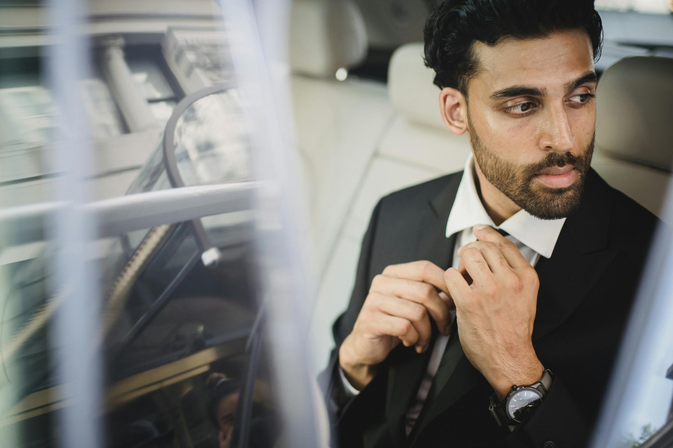Bearded man in black suit sitting in car adjusting tie with serious expression.