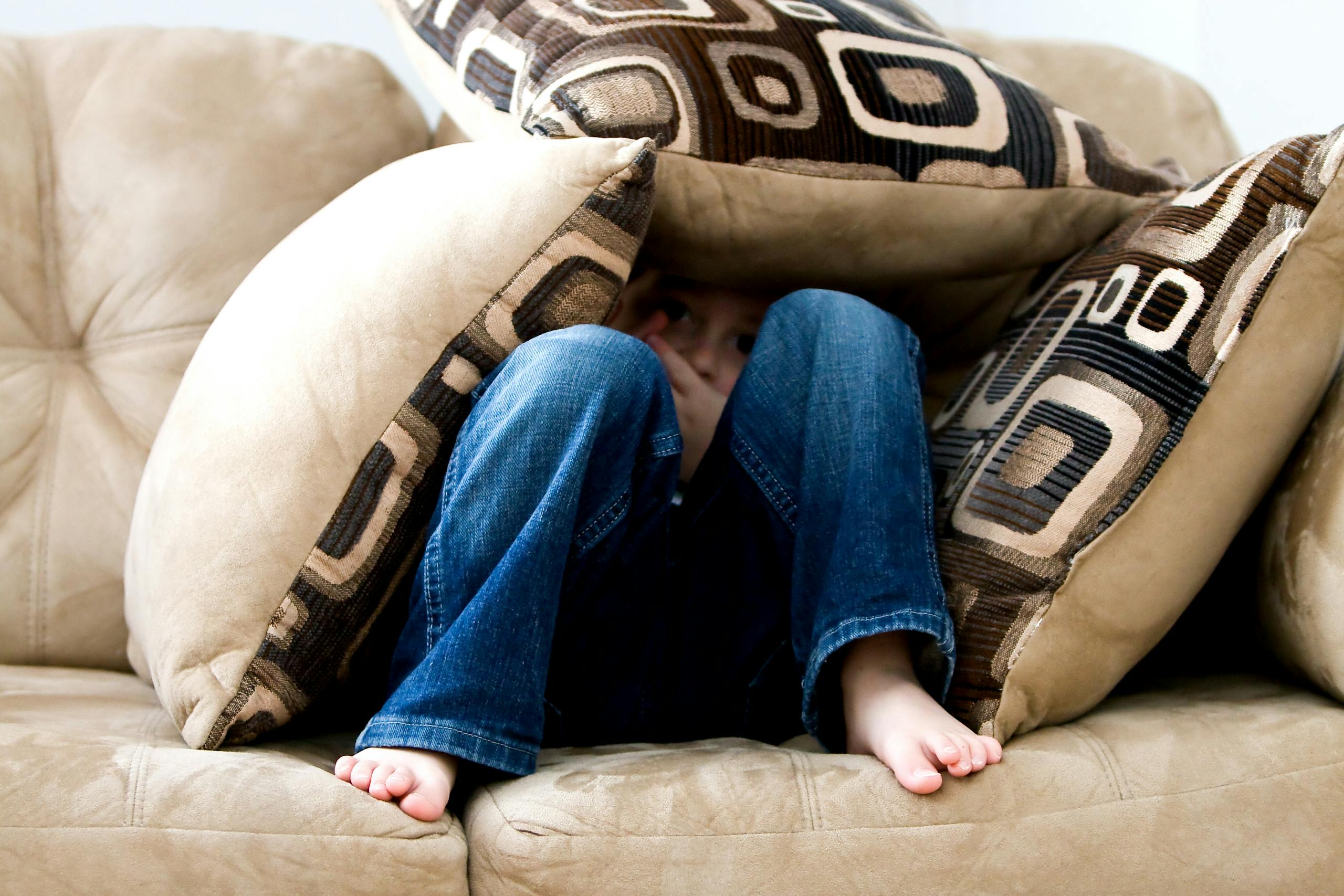 Child crouched under cushions on couch in playful or fearful moment.