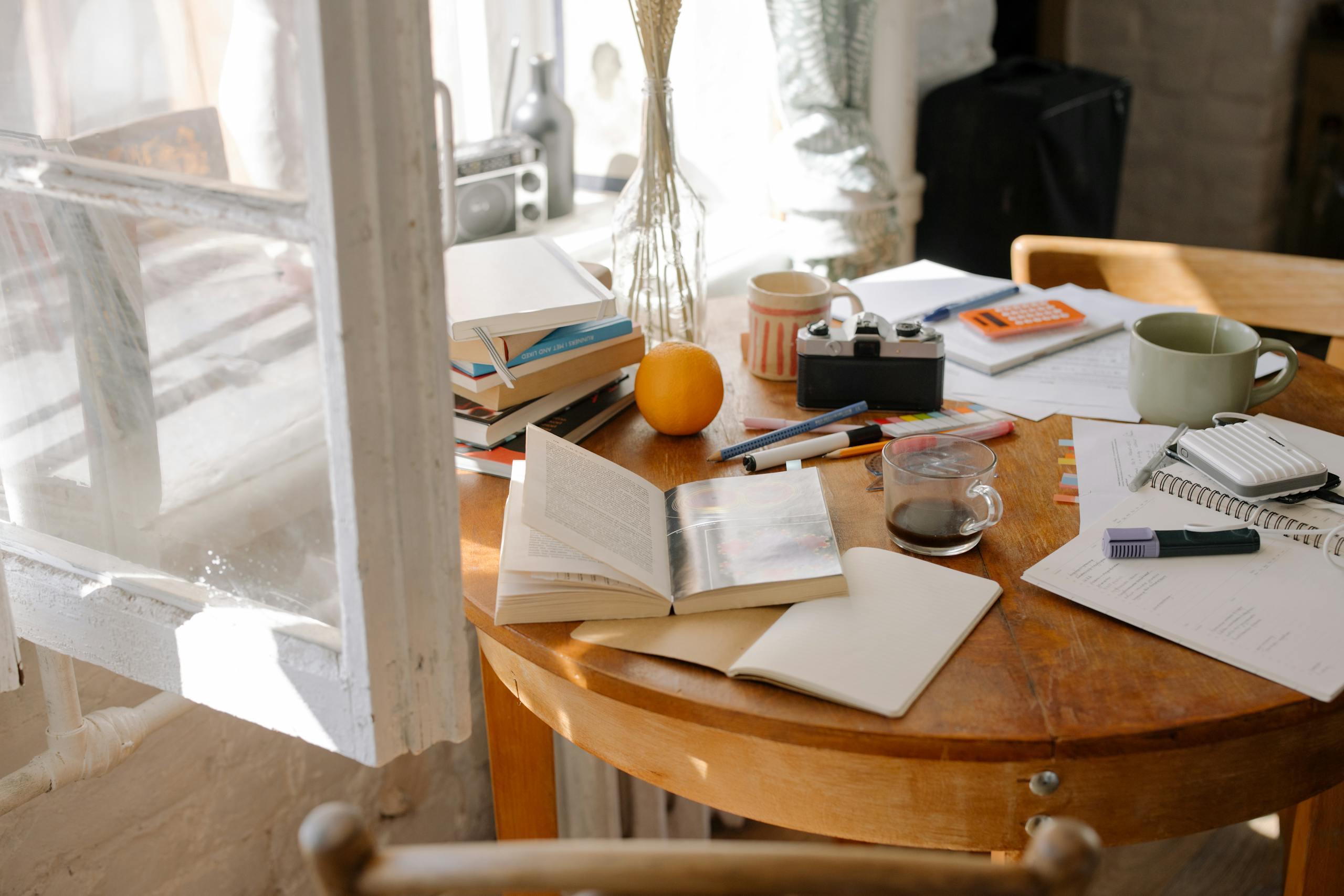 Cluttered study desk with open books, coffee, and sunlight streaming through window.