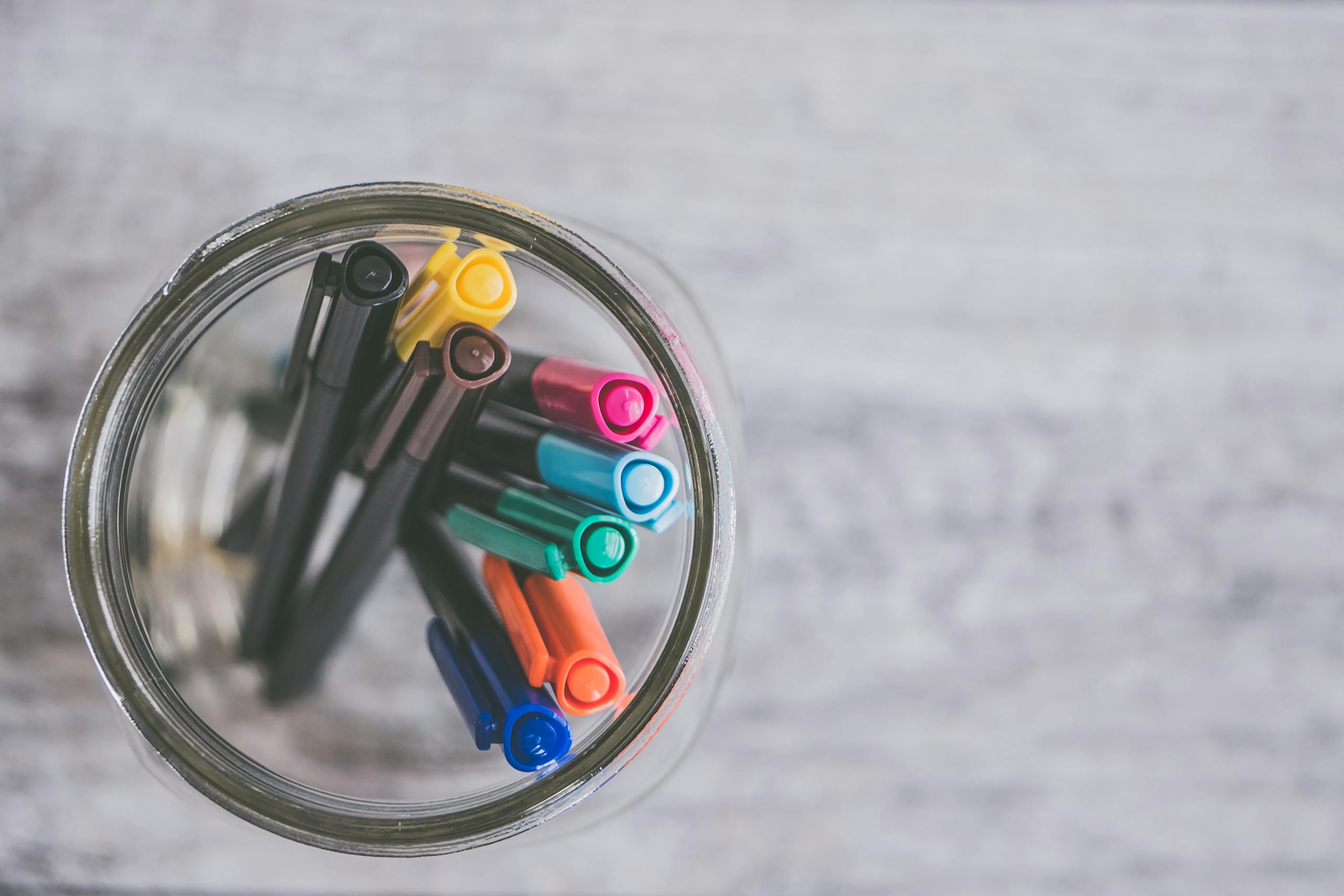 Flat lay of colorful pens arranged in mason jar on light wooden desk.