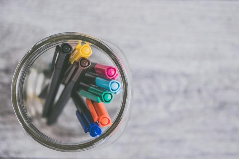 Flat lay of colorful pens arranged in mason jar on light wooden desk.