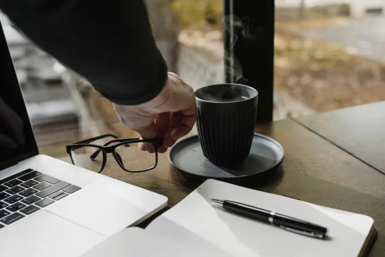 Hand reaches for steaming coffee cup near laptop and open notebook in cozy workspace