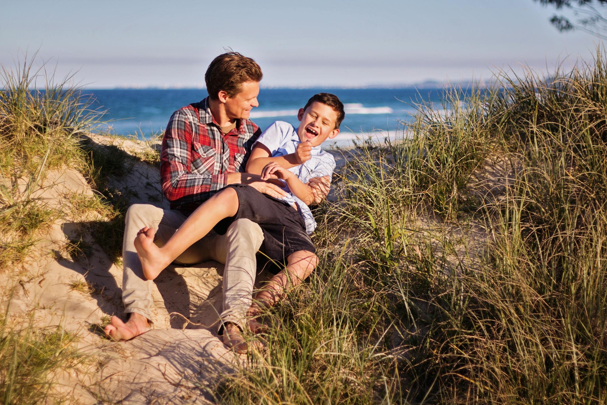 Joyful father and son share laugh on sunny beach surrounded by sand dunes