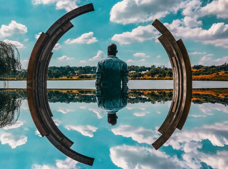 Man in denim jacket sitting contemplatively by abstract sculpture and water.