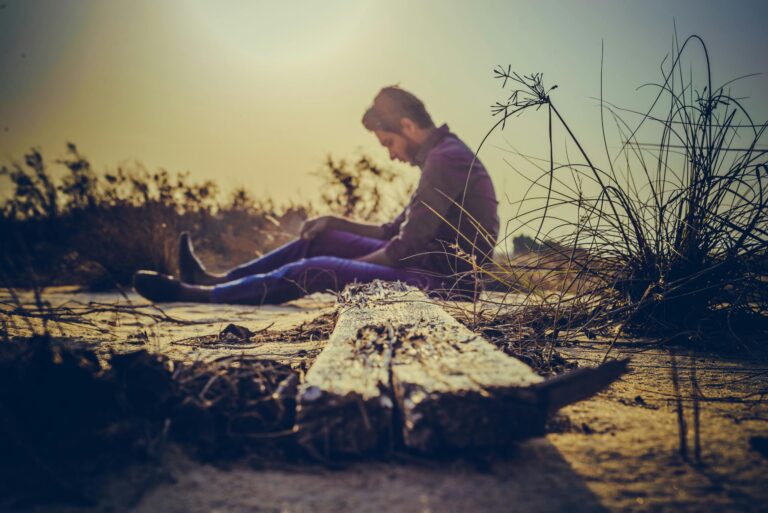 Man sits alone on sandy terrain at sunset capturing solitude in nature's calm.