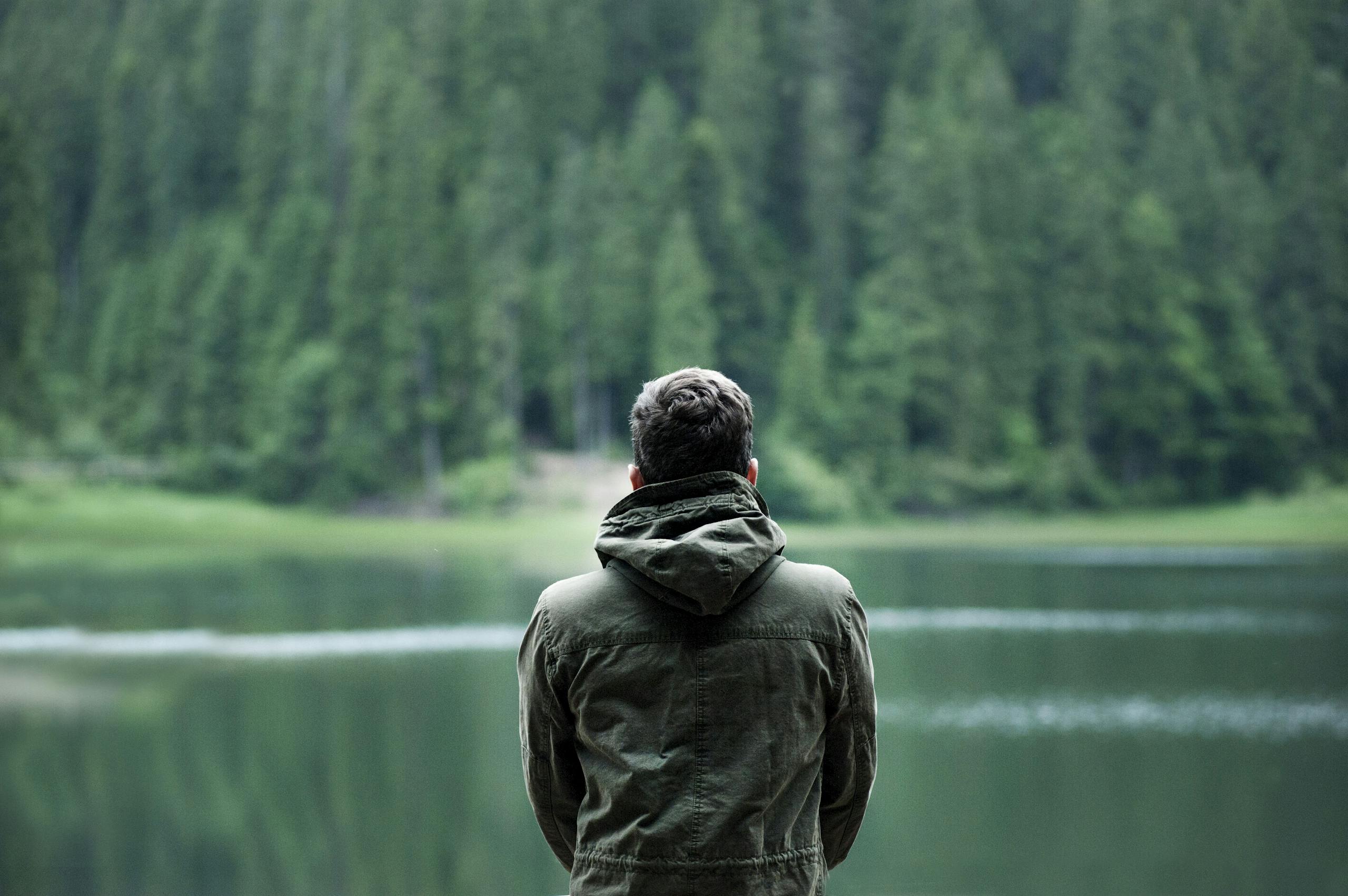 Person in green jacket standing by serene forest lake in calming natural setting