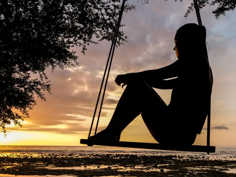 Woman silhouetted on swing by seashore at sunset evoking tranquility.