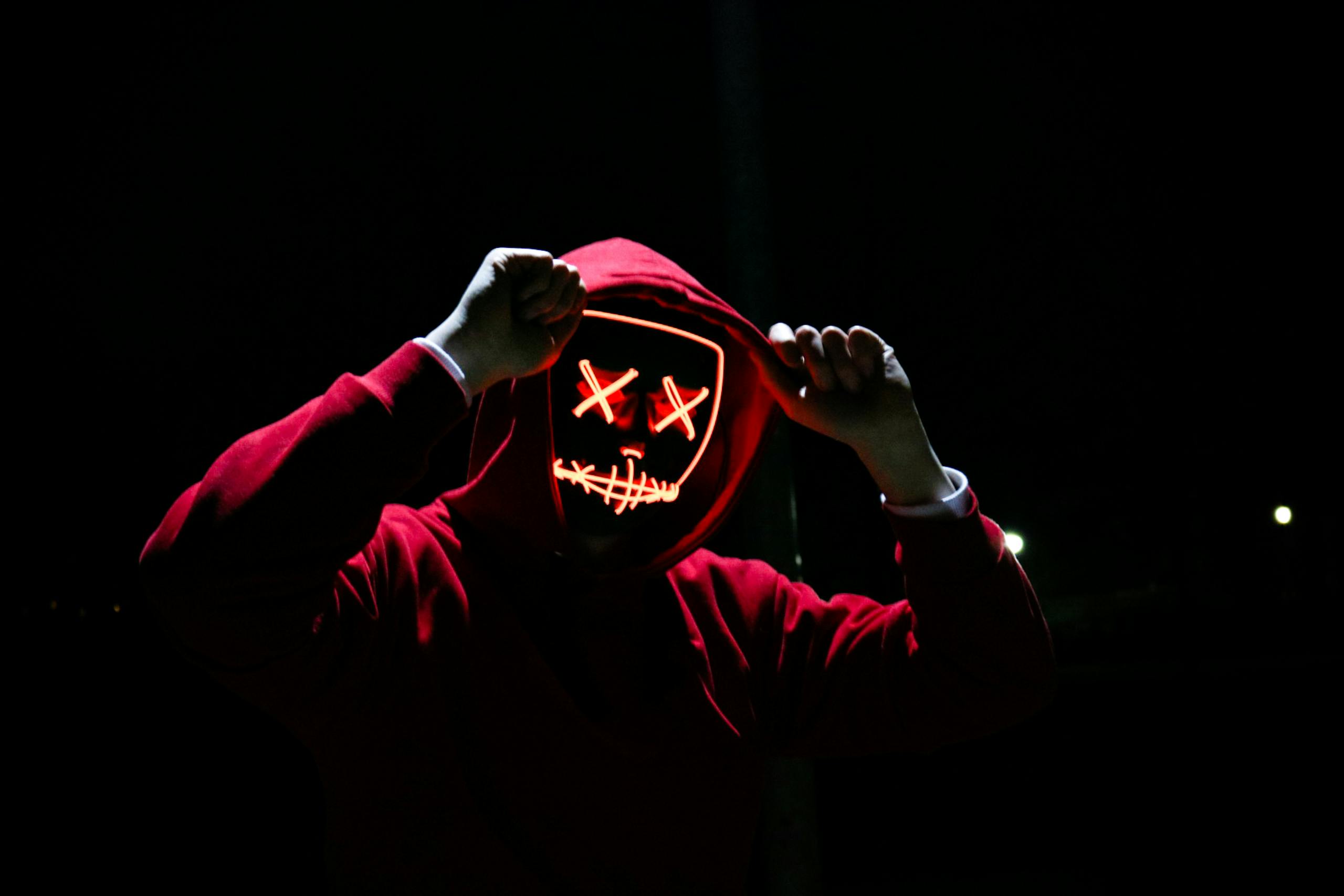 Spooky illuminated mask figure wearing red hoodie in darkness