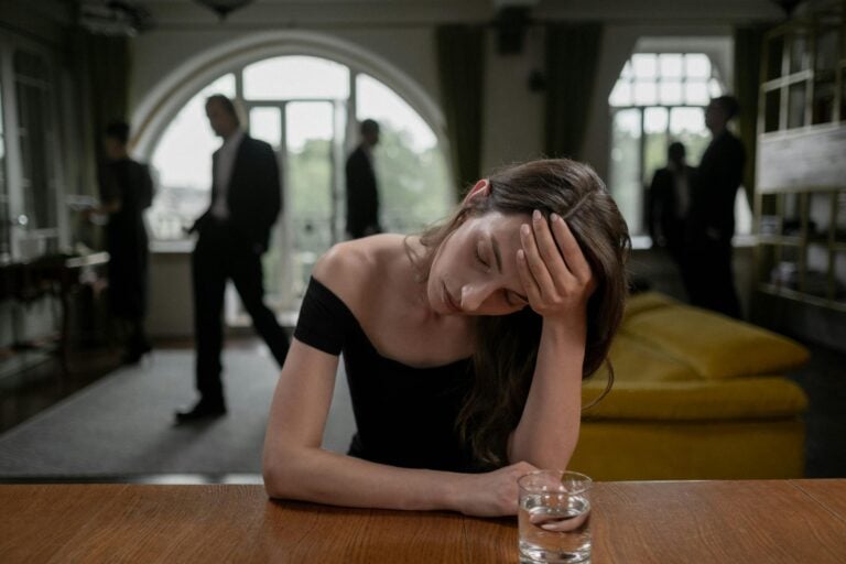 Stressed woman in formal indoor setting with glass of water and blurred background figures