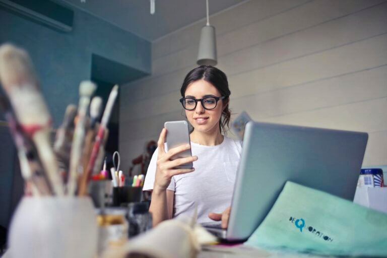 Woman using phone at desk surrounded by art supplies and laptop.