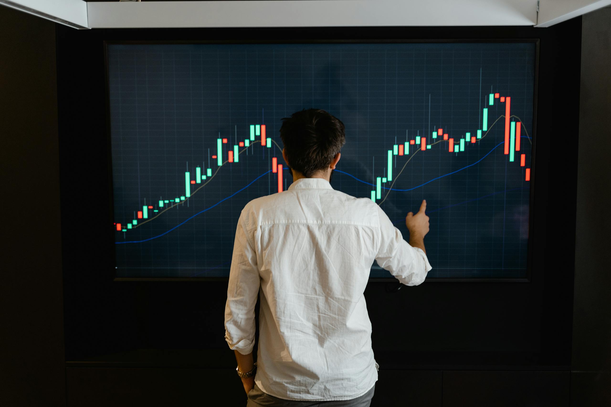 Adult male reviewing stock market data on large display screen indoors.